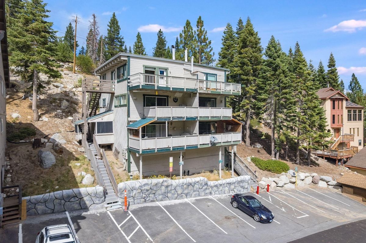 A multi-story building with a balcony, a parking area, and a mountainous backdrop.