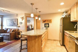 A kitchen with a central island, wooden cabinets, a refrigerator, and a dining area with a sofa and a coffee table.