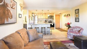 A living room with a brown leather couch, a patterned armchair, and a kitchen area with wooden cabinets and a bar stool.