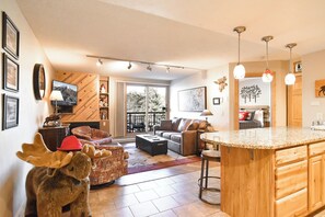 A modern living room with a fireplace, a sofa, a coffee table, and a kitchen area with a wooden countertop and bar stools. - Trails End Moose Haven Condo - Ski In/Out, Walk to Town! (Breckenridge)