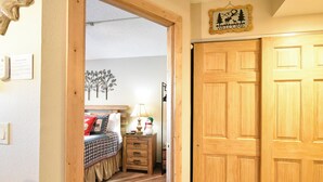 A wooden door with a 'Welcome' sign above it, a bed with a checkered bedspread, a nightstand with a lamp, and a wall with a tree decoration.