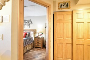 A wooden door with a 'Welcome' sign above it, a bed with a checkered bedspread, a nightstand with a lamp, and a wall with a tree decoration.