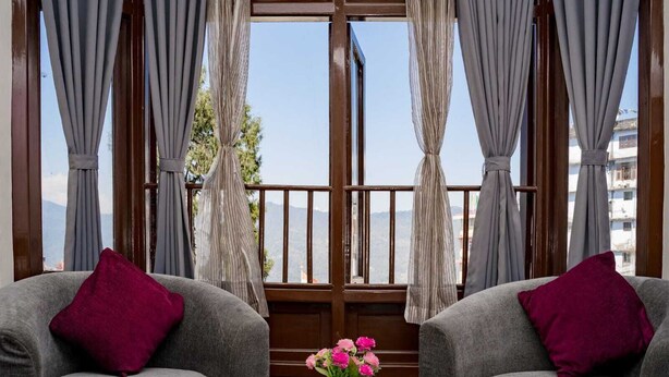 Una habitación con dos sillones grises, una mesa de madera y vistas a montañas a través de un gran ventanal con cortinas.