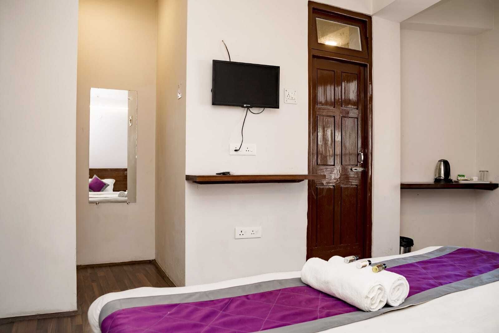 Una habitación de hotel con una cama, un televisor montado en la pared, una puerta de madera y un estante con una tetera y tazas.