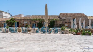 A courtyard with stone flooring, outdoor seating with blue chairs, and a building with a tiled roof.