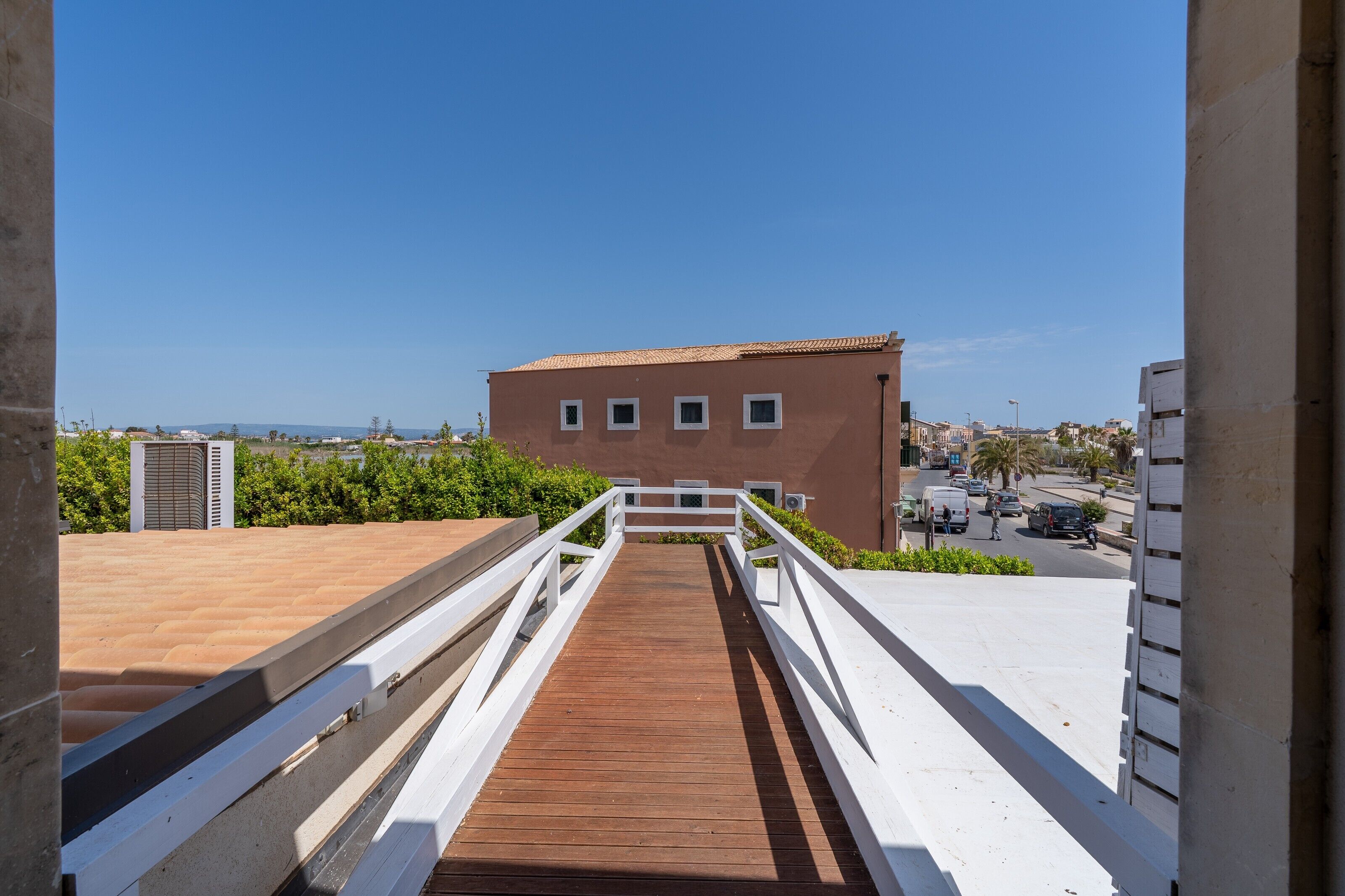 A wooden deck leading to a building with a red-tiled roof, surrounded by greenery and a clear blue sky.