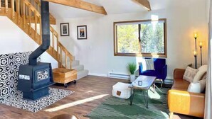 Un salon moderne avec un escalier en bois, un poêle à bois encastré, un canapé, une table basse et une grande fenêtre donnant sur la forêt.
