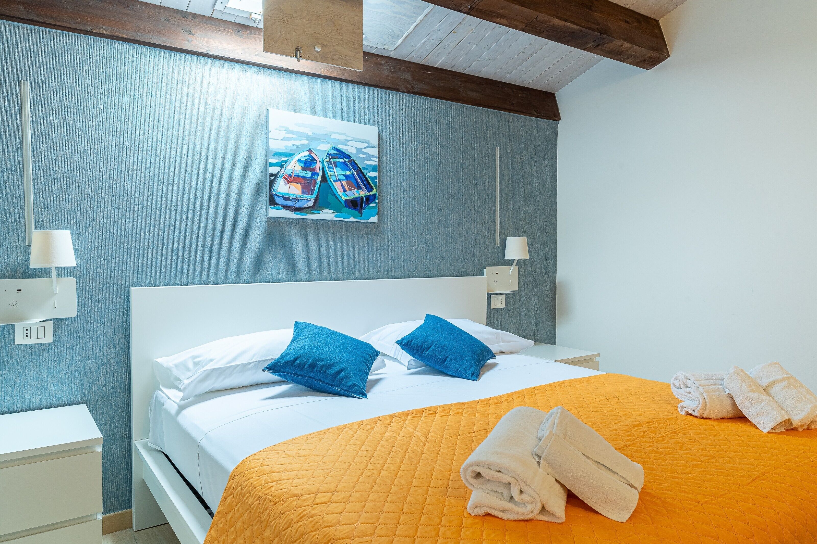 A hotel room with a bed, two pillows, a painting, a nightstand, and a folded towel.