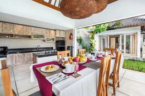 Una cucina moderna con un'area pranzo. La cucina presenta un piano di lavoro nero, un lavello e mobili in legno. L'area pranzo include un tavolo apparecchiato con piatti, tazze e una ciotola di frutta. Sopra il tavolo, un lampadario in vimini.