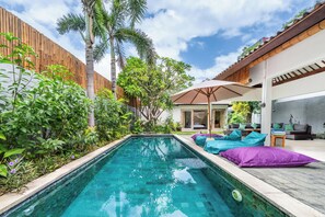 Una piscina circondata da vegetazione lussureggiante, un recinto in legno e un'area salotto esterna moderna con cuscini.