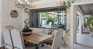 A dining area with a wooden table, white upholstered chairs, a large window, and a decorative mirror.