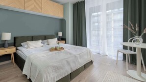 Ein modernes Schlafzimmer mit einem großen Bett, Nachttischen, einem kleinen Tisch mit einer Vase und Ausblick ins Freie durch transparente Vorhänge.