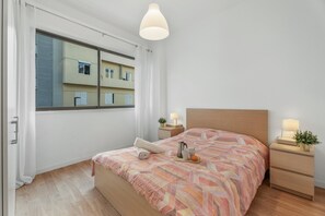 Un dormitorio con cama, mesitas de noche, ventana y vistas a edificios.