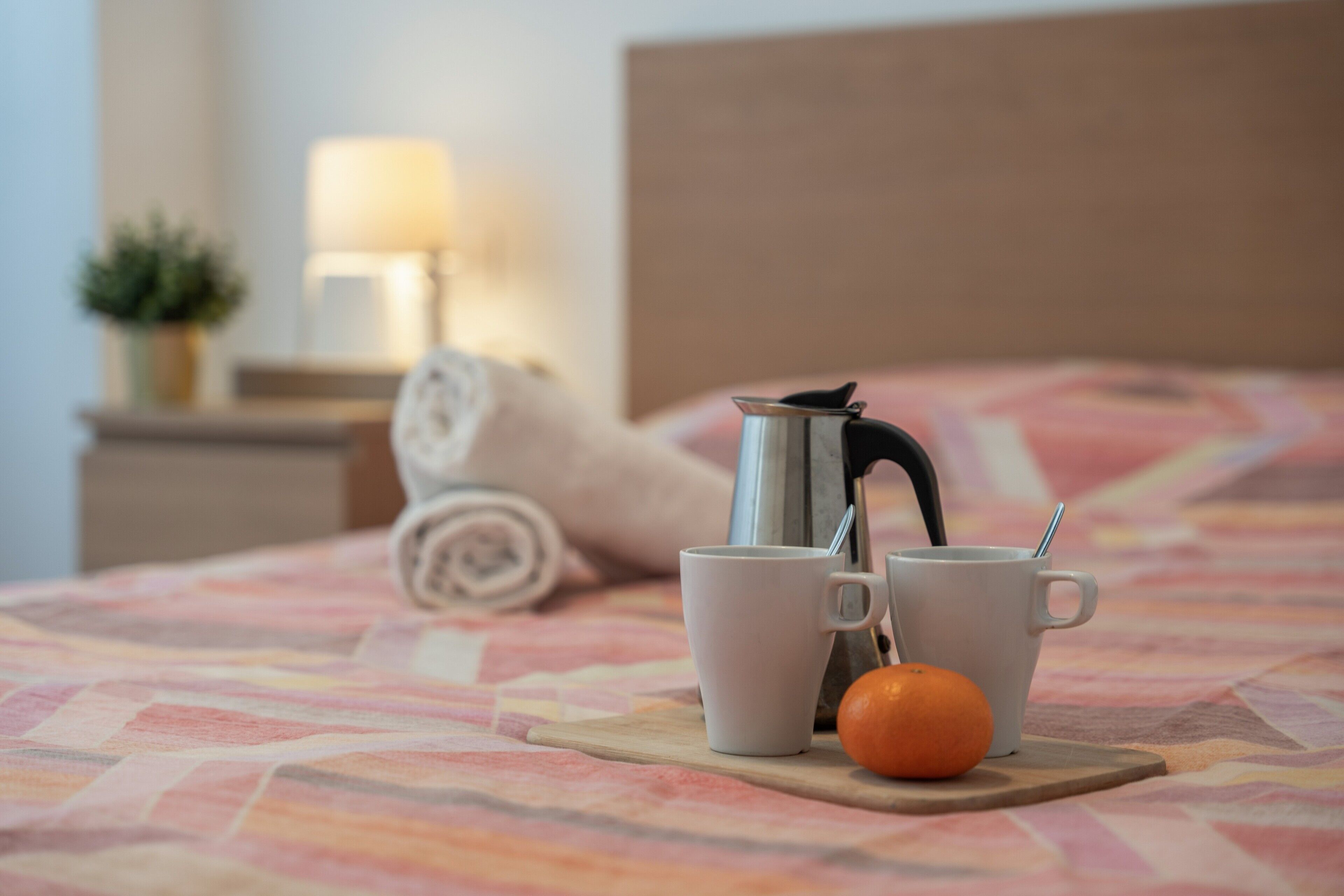 Ein Bett mit gemustertem Bettbezug, ein Nachttisch mit Lampe, ein zusammengerolltes Handtuch und zwei Tassen mit Teekanne und einer Orange auf einem Holztisch.