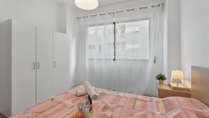 A bedroom with a bed, a bedside table with a lamp, a window with sheer curtains, and a wardrobe.