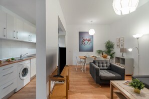 A modern kitchen with a washing machine, a dining area with a table and chairs, a grey sofa, and a ceiling light fixture.