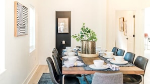 A dining area with a wooden table set for six, surrounded by black chairs. A large potted plant is centered on the table.