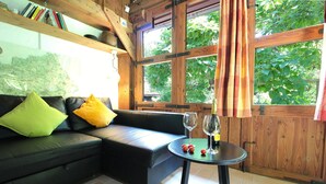 Une pièce chaleureuse avec un canapé en cuir noir, une petite table ronde sur laquelle se trouvent un verre de vin et des tomates, et de grandes fenêtres avec des rideaux.