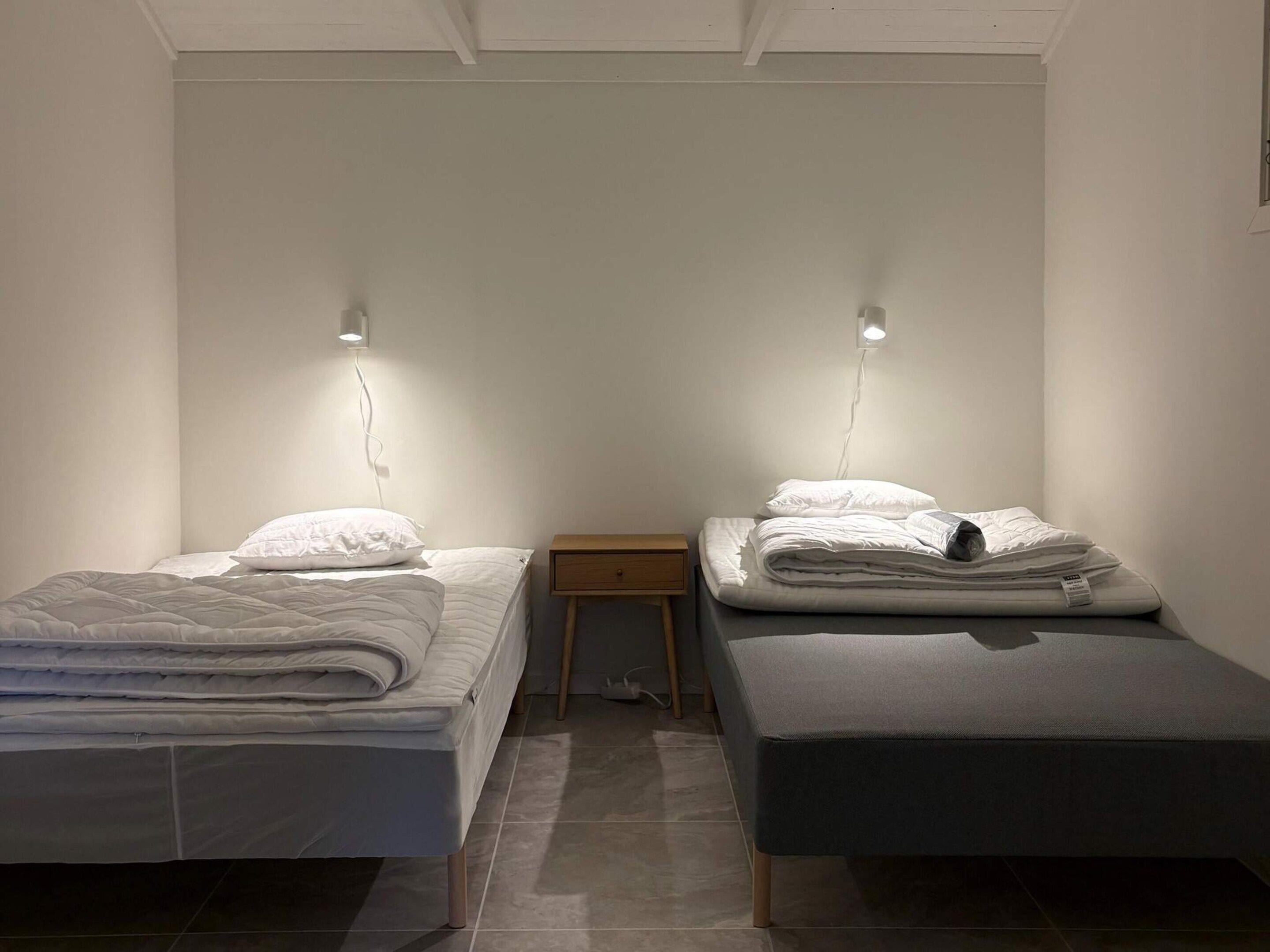 Una habitación con dos camas individuales, cada una con una almohada y una manta doblada. Hay una mesita de noche de madera entre las camas.