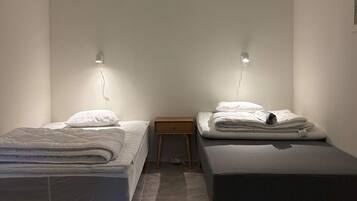 Una habitación con dos camas individuales, cada una con una almohada y una manta doblada. Hay una mesita de noche de madera entre las camas.