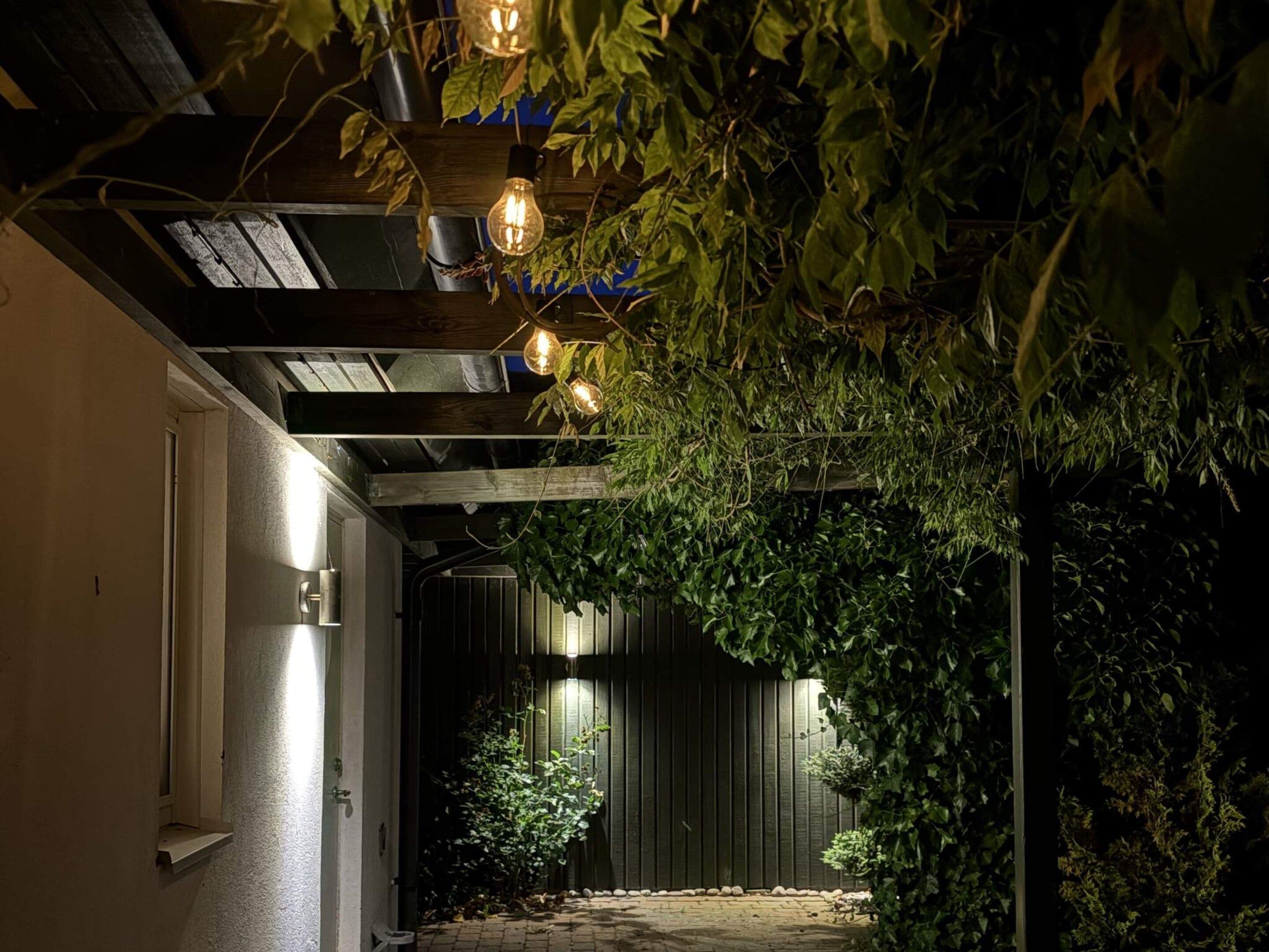 An outdoor corridor with a wooden ceiling, hanging light bulbs, and a dark background.