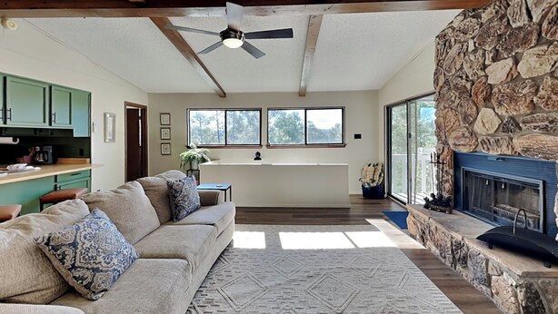 A spacious living room with a stone fireplace, a built-in kitchen with green cabinets, and a large window overlooking a forested area. - Fireside Cabin | Close to Midtown + Trails (Ruidoso)