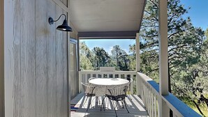 A covered outdoor deck with a round table and chairs, a white railing, and a view of trees and mountains in the background. - Fireside Cabin | Close to Midtown + Trails (Ruidoso)
