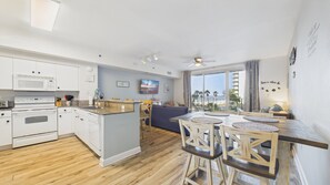 A modern kitchen with white appliances, a dining area with wooden chairs, and a view of the beach.