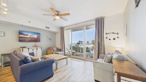 A living room with a sofa, a coffee table, and a TV mounted on the wall. There is a balcony with a view of the beach.