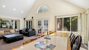 Ein geräumiges Wohnzimmer mit hohen Decken, großen Fenstern, einem Essbereich mit Tisch und Stühlen sowie einer gemütlichen Sitzecke mit Sofa und Hocker.