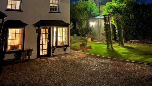 Ein zweistöckiges Haus mit mehreren erleuchteten Fenstern, einem gepflegten Rasen und einem sichtbaren Gartenbereich.