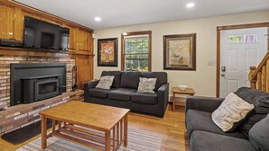 A living room with a fireplace, a sofa, a coffee table, and framed pictures on the wall. - Cape Cod Family Escape w/ Pool, Game Room & Patio (Falmouth)
