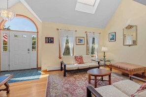 A spacious living room with a sofa, a round table, a bench, and a large window with curtains.