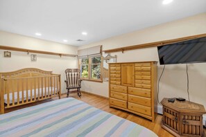 A bedroom with a wooden crib, a wooden dresser, a television, and a window with a view of greenery. - Bright & Spacious Poolside Cape Getaway (Mashpee)