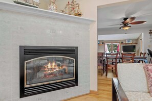 A living room with a fireplace, a ceiling fan, and a dining area in the background.