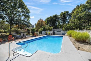 A rectangular swimming pool with a metal ladder, surrounded by a concrete deck, trees, and a fence.