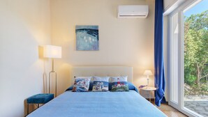A bedroom with a bed, a painting, an air conditioner, a lamp, and a view of greenery.
