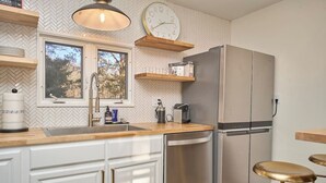 Ett modernt kök med vita skåp, ett kylskåp i rostfritt stål, bänkskivor i trä och en inbyggd ugn. Det finns två fönster som släpper in naturligt ljus och en klocka på väggen.