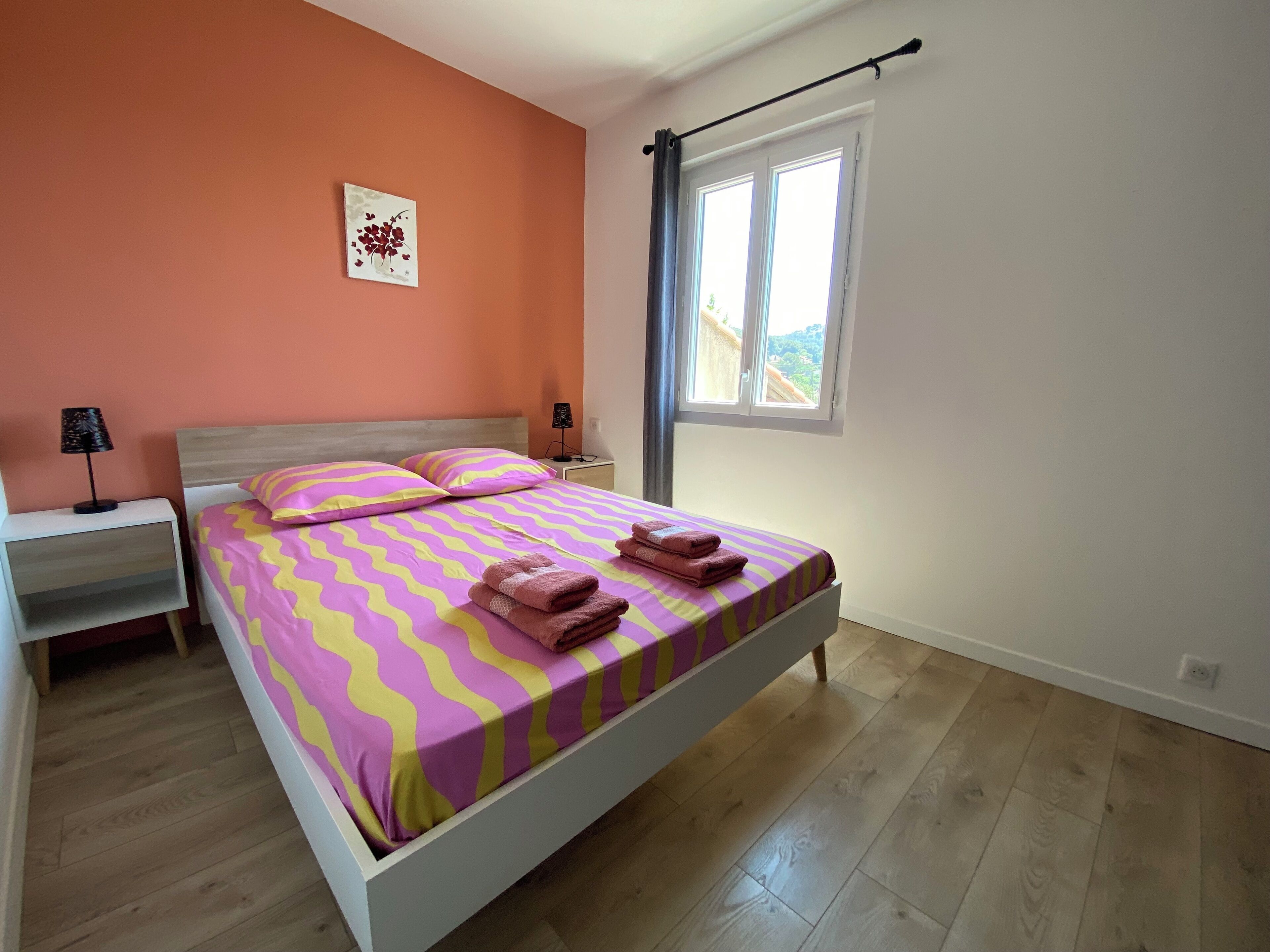 Ein Schlafzimmer mit einem Bett, einem Nachttisch, einer Lampe, einem Fenster mit Vorhängen und einem an der Wand hängenden Kunstwerk.