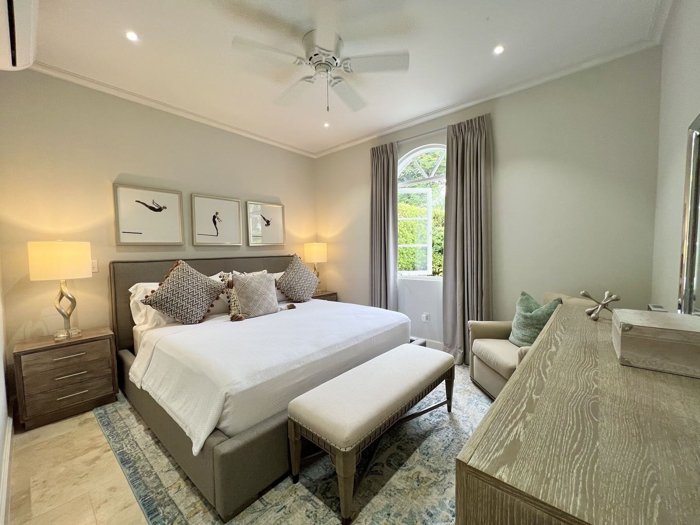A bedroom with a bed, bedside tables, a bench, a chair, a ceiling fan, and a window with curtains.