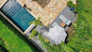 Luchtbeeld van een zwembad met betegeld zonnedek, omgeven door weelderige begroeiing en een houten terras met een witte parasol.