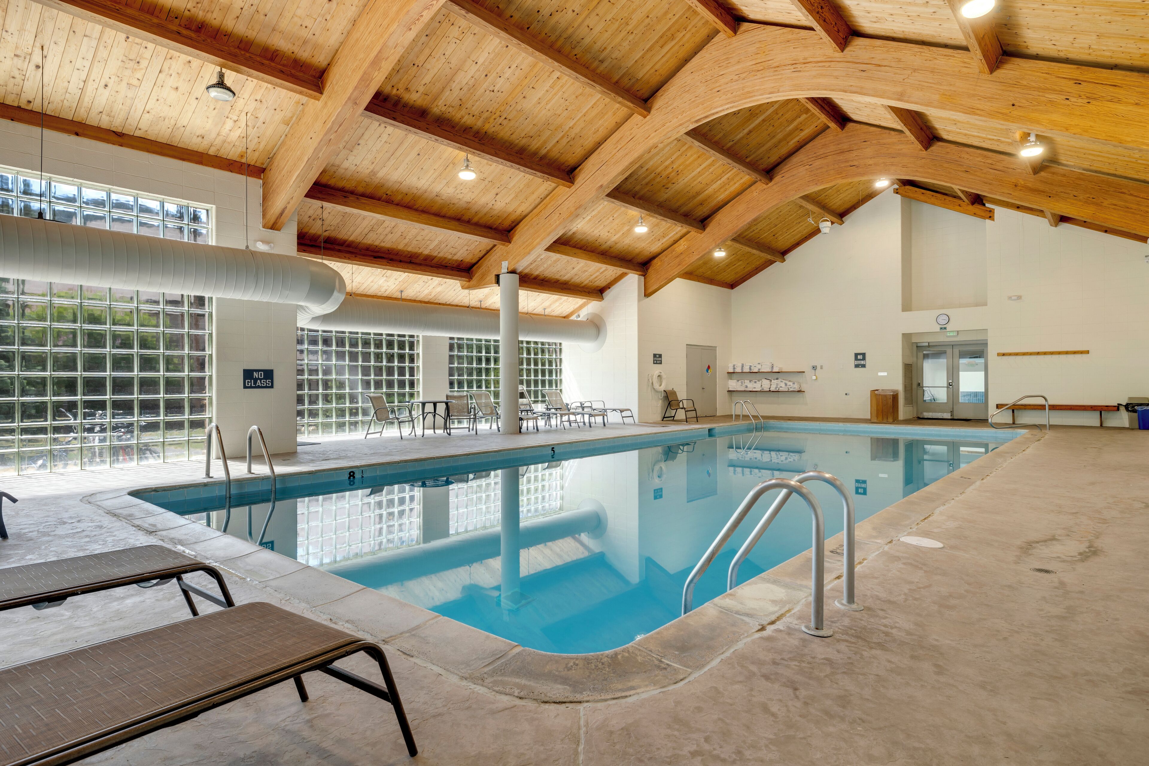An indoor swimming pool with a wooden ceiling, clear glass windows, and lounge chairs.