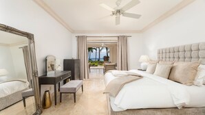 A spacious bedroom with a large bed, a vanity with a mirror, a desk, and a view of the ocean through an open door. - St. Peter's Bay 111 2 Bedroom Condo (Speightstown)