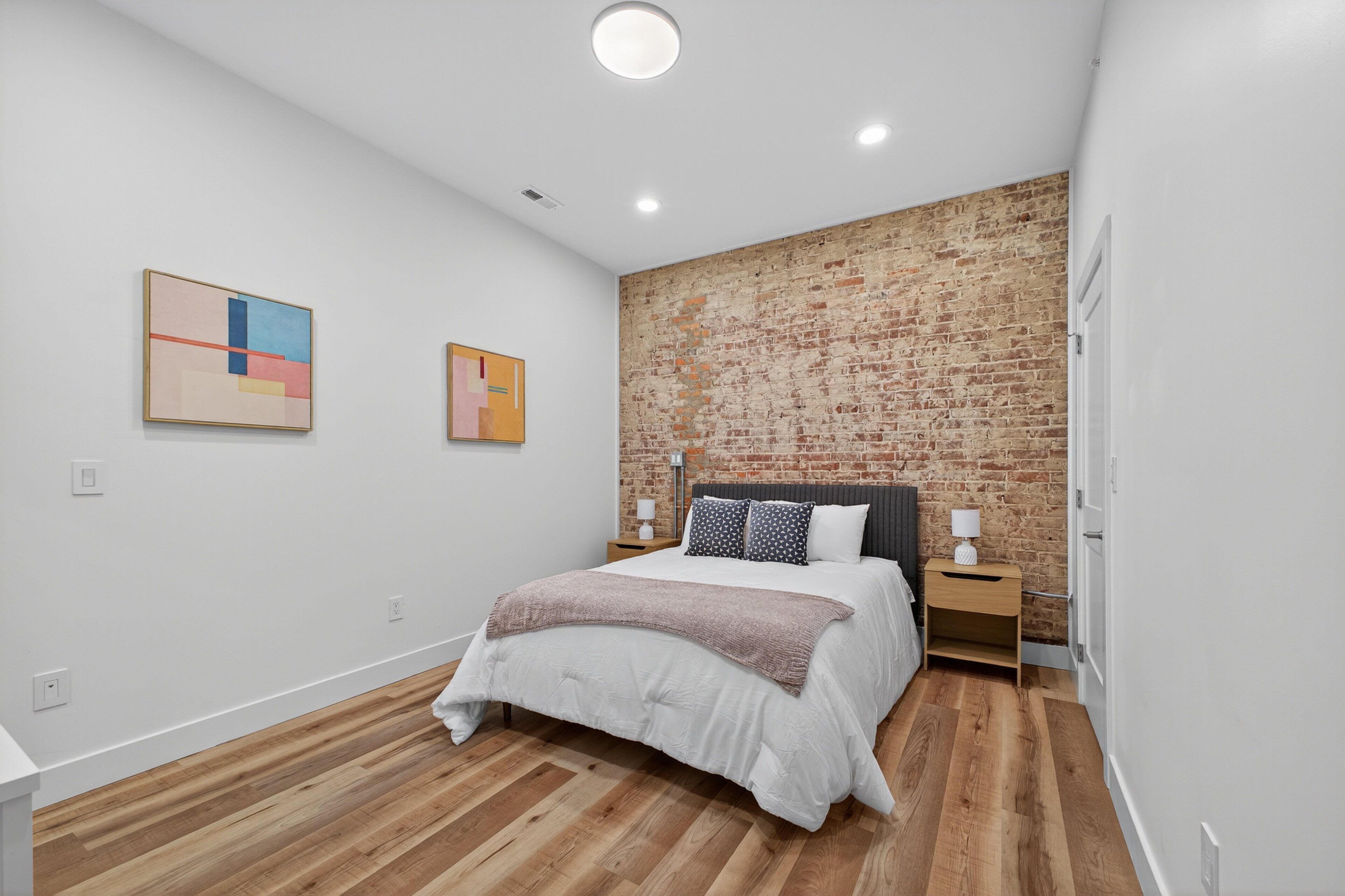A modern bedroom with a brick accent wall, a large bed with a white comforter, two bedside tables with lamps, and abstract artwork on the walls.