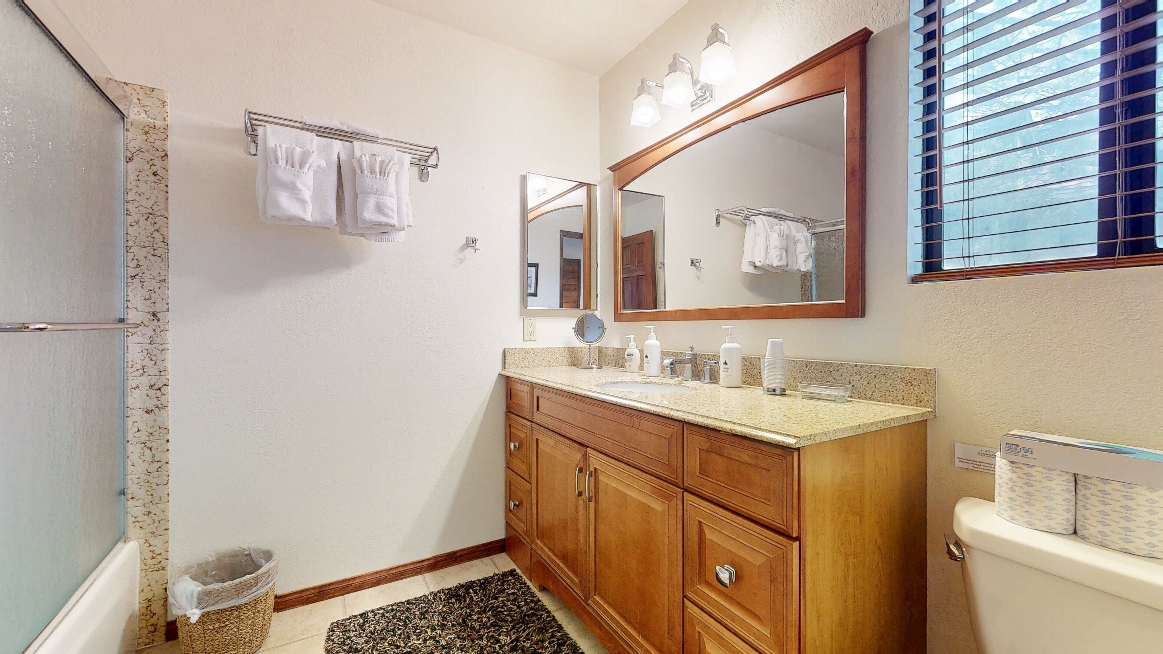 A bathroom with a wooden vanity, a large mirror, and a toilet. There are towels hanging on a rack and a small trash bin.