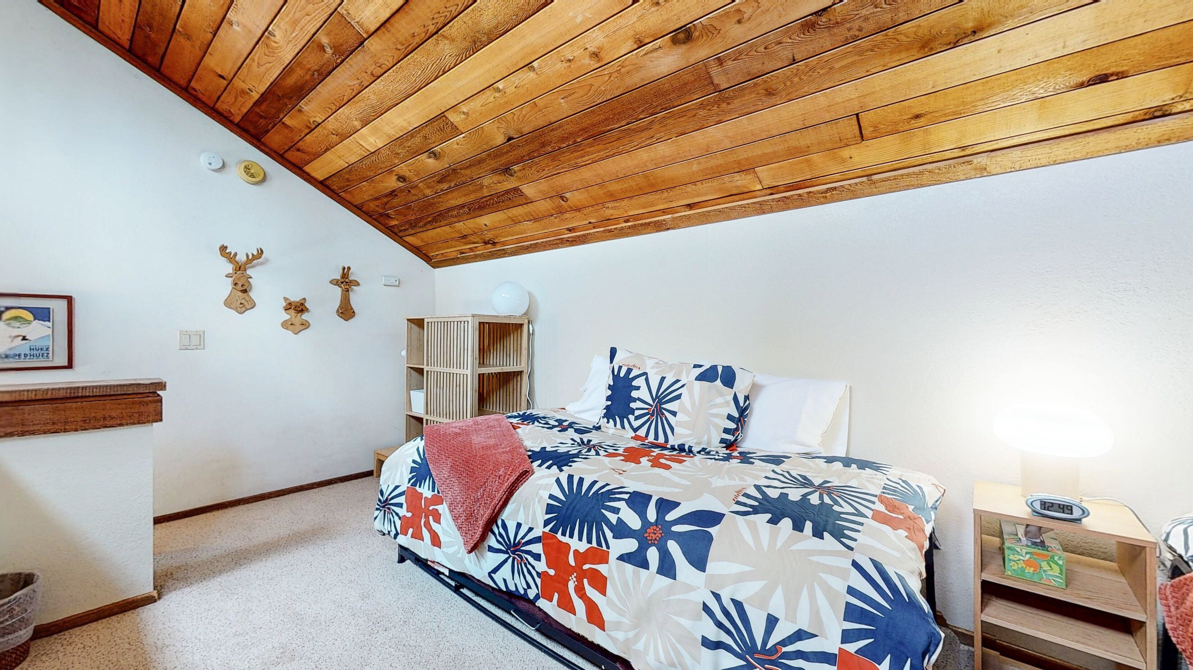 Une chambre avec un plafond en bois, un lit recouvert d’une couvre-lit à motifs floraux, une table de chevet avec une horloge et une étagère ornée d’objets décoratifs.