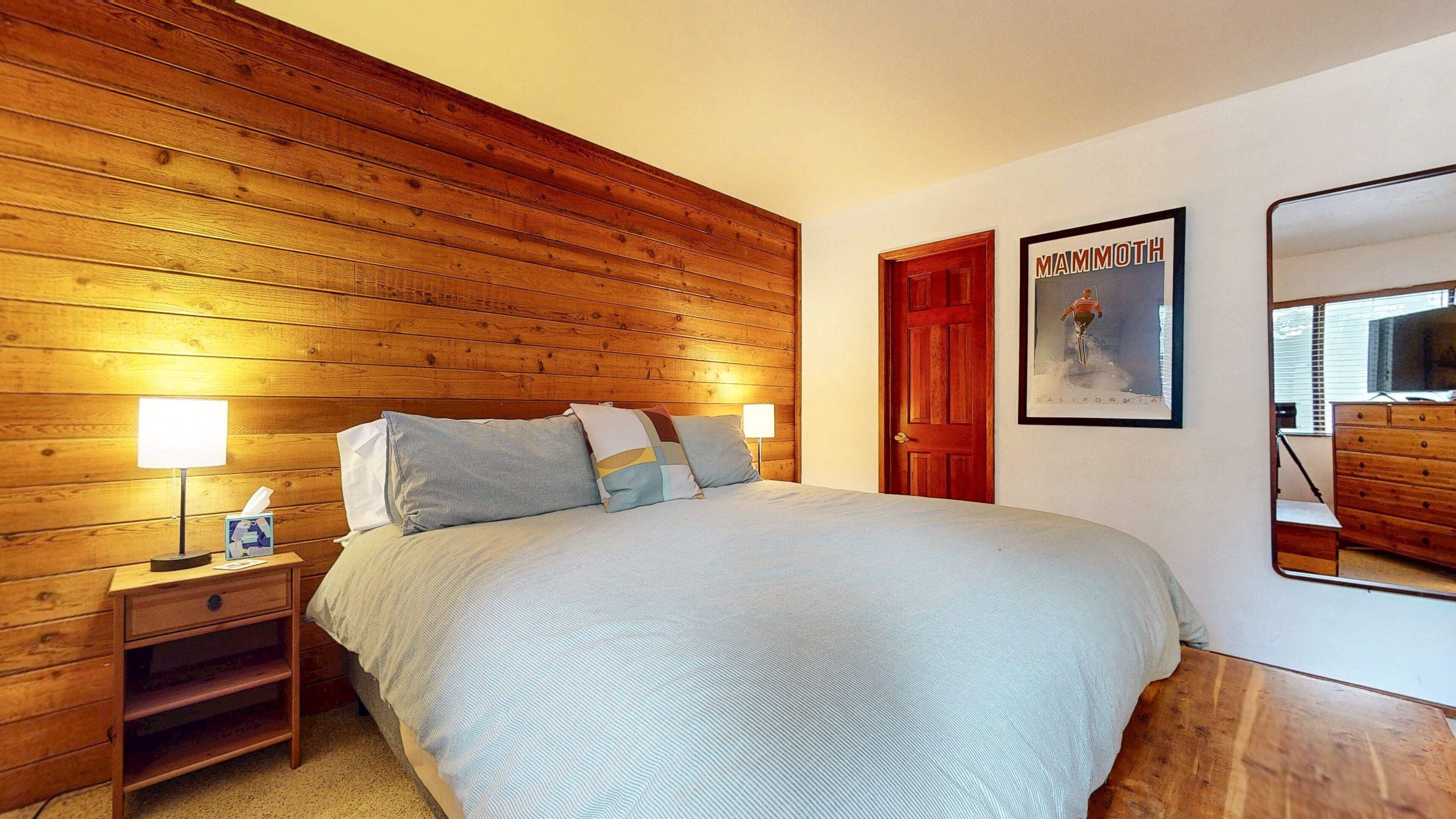 A bedroom with a wooden headboard, a bed with a striped bedspread, two bedside lamps, a nightstand with a tissue box, and a framed picture on the wall.