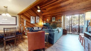 A cozy cabin interior with wooden walls, a stone fireplace, a television, and a dining area with a table and chairs. - Mammoth Sierra Townhomes #25 3 Bedroom Condo (Mammoth Lakes)