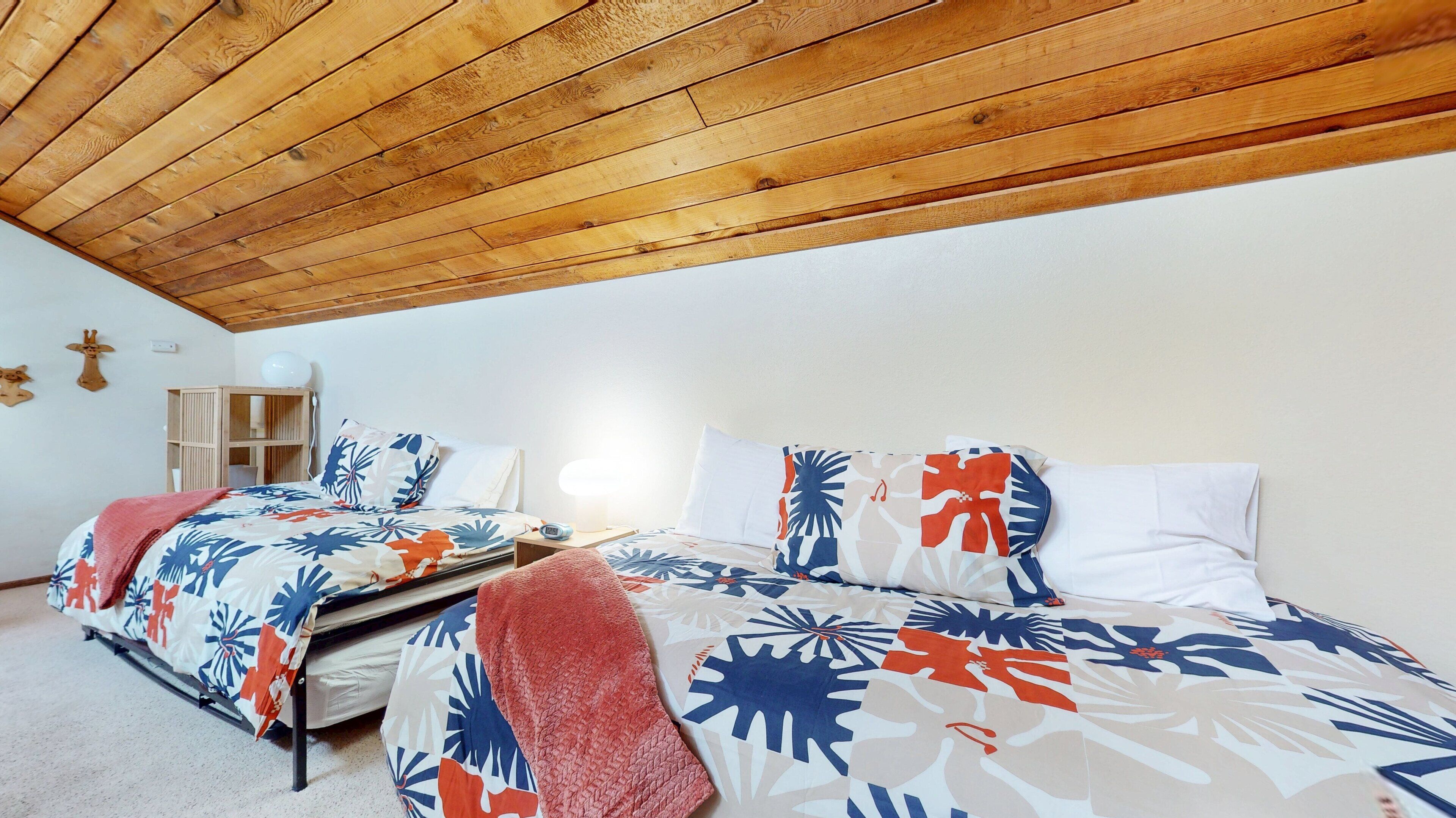 Une chambre avec un plafond en bois, un lit recouvert d’une literie à motifs floraux, une table de chevet et une petite table de nuit.