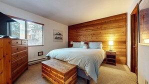 A bedroom with a wooden headboard, a bed with multiple pillows, a nightstand with a lamp, a wooden dresser, and a window with blinds. - Mammoth Sierra Townhomes #25 3 Bedroom Condo (Mammoth Lakes)